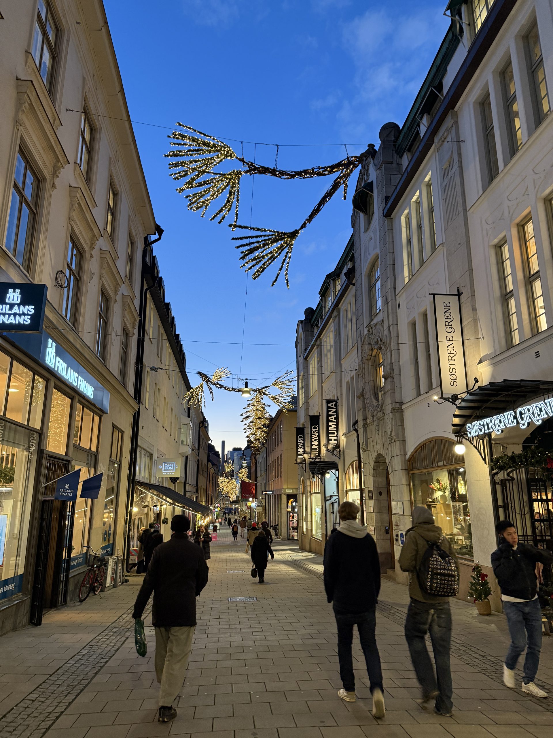 Människor promenerar på en gata med upplysta butiker och julbelysning.