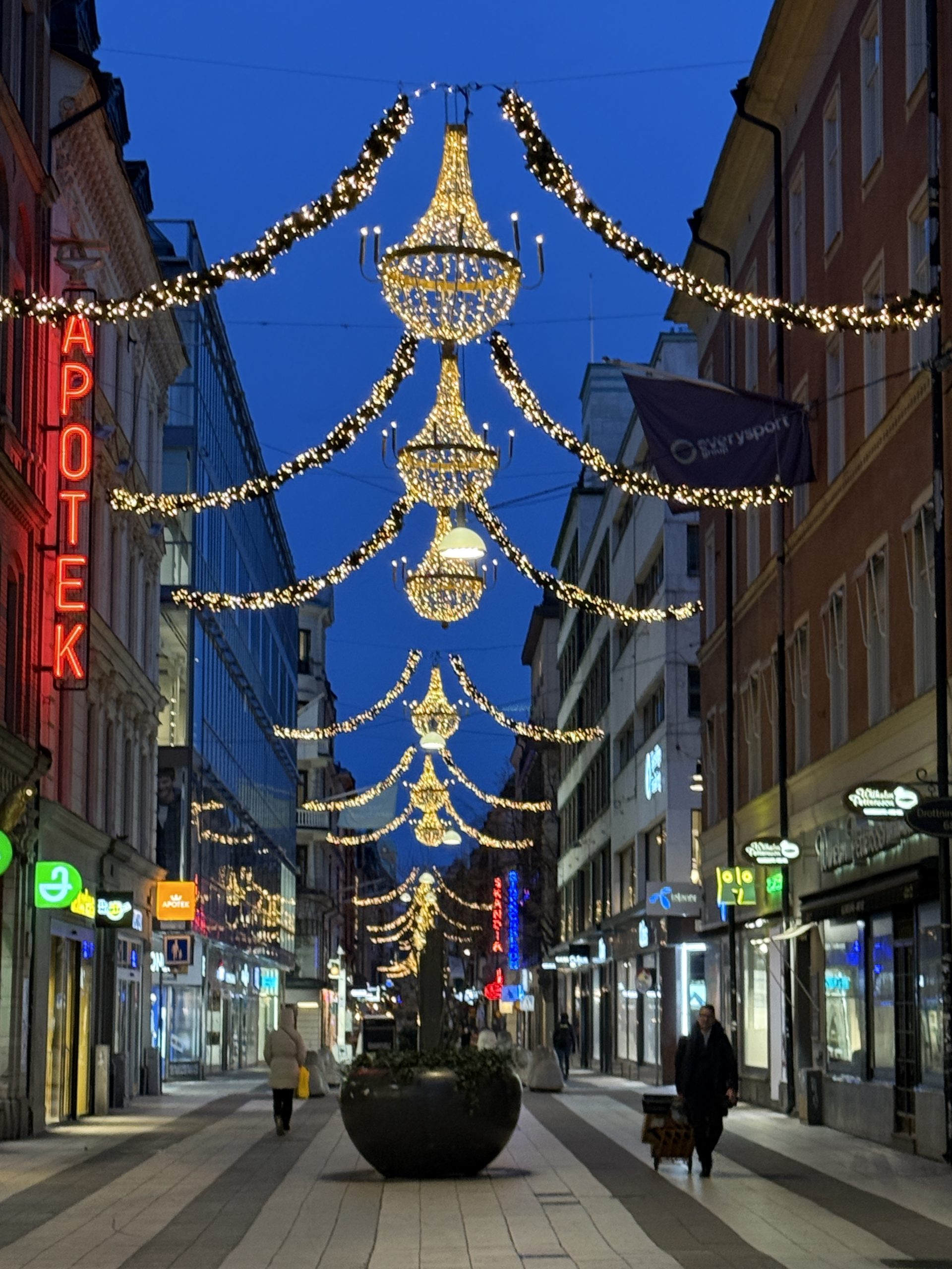 Julbelysning hängandes mellan fastigheter på Drottninggatan i Stockholm. Kvällstid med blå himmel.
